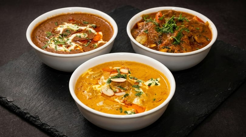Three bowls of Indian curries, two brown and one orange, garnished with cream, herbs, and nuts.