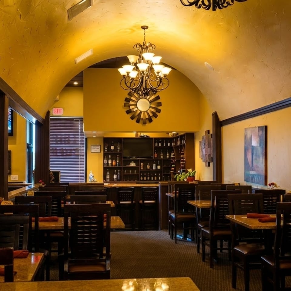 Warmly lit restaurant interior with yellow walls, dark wood tables, chairs, and a bar area.